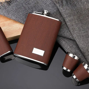 Elegant stainless steel hip flask set with a wooden finish and matching shot cups on a textured backdrop.