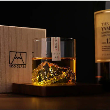 Japan 3D Mountain Whisky Glass with whisky on the rocks, showcasing a stunning 3D mountain design, placed on a wooden coaster next to a bottle and box.