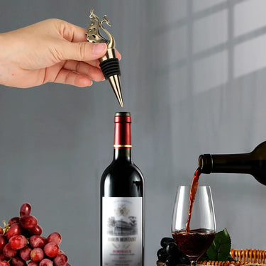 Swan wine stopper being placed on a wine bottle next to a glass being poured with red wine and a bunch of grapes.