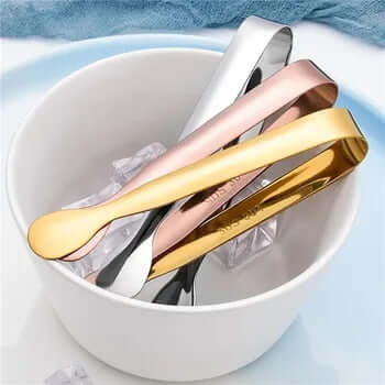 Stainless steel ice cube tongs in silver, rose gold, and gold resting in a bowl of ice cubes. Perfect for drink service.